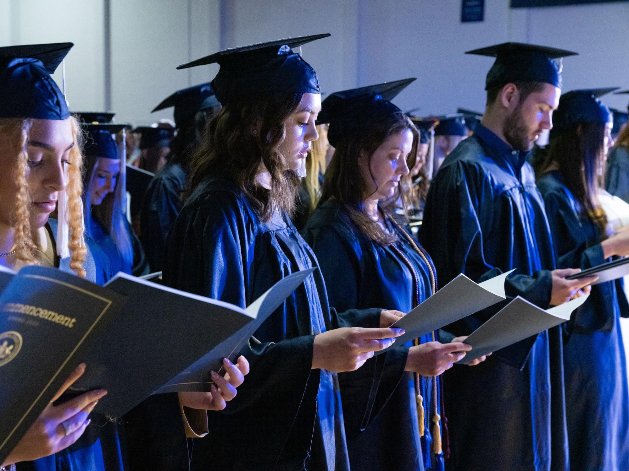 Students in caps and gowns reading a booklet during graduation.
