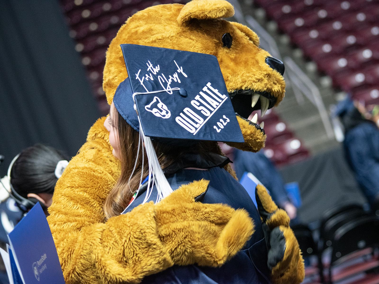 The Nittany Lion hugs a graduate in a cap that reads "For the Glory of Old State 2023"