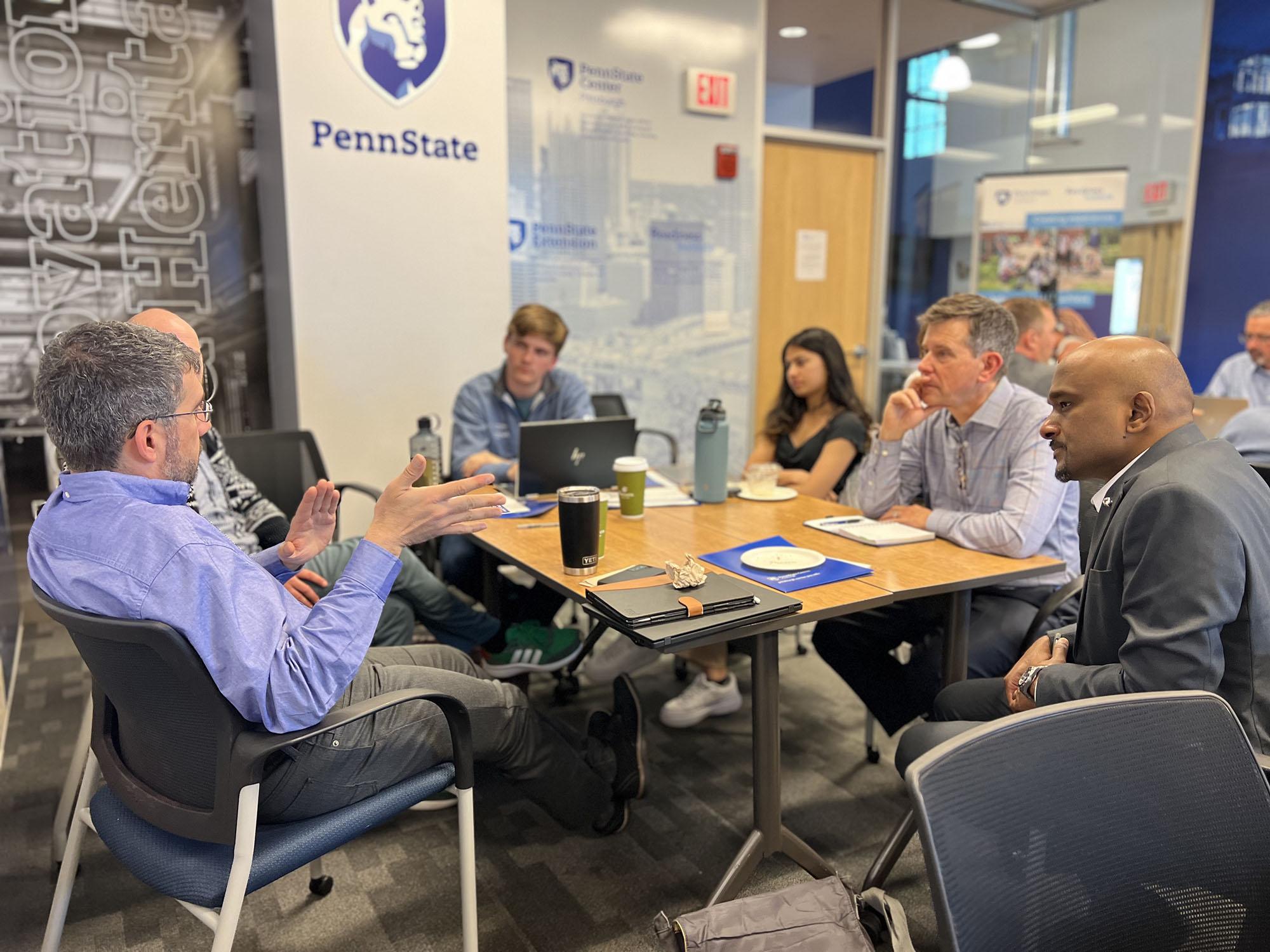 A group of people sit around a table having a discussion. A Penn State logo is on a wall in the background. 