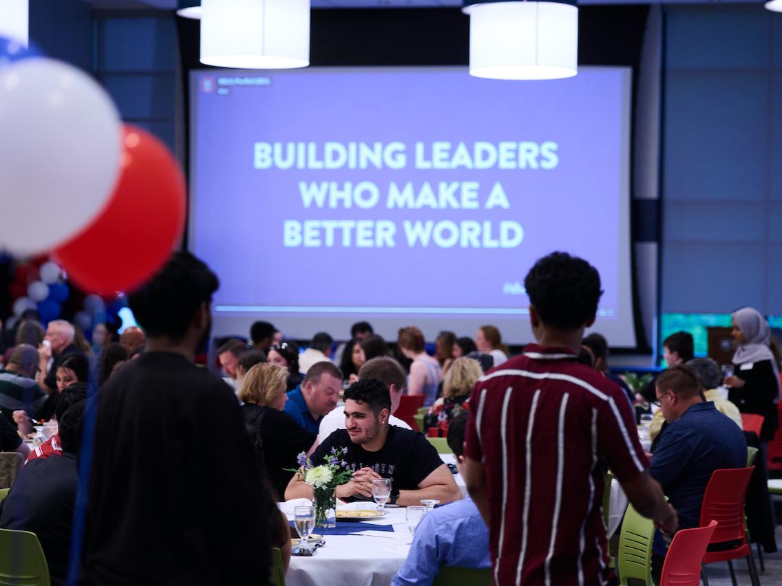 crowd sitting at tables with "building leaders who make a better world" on projector