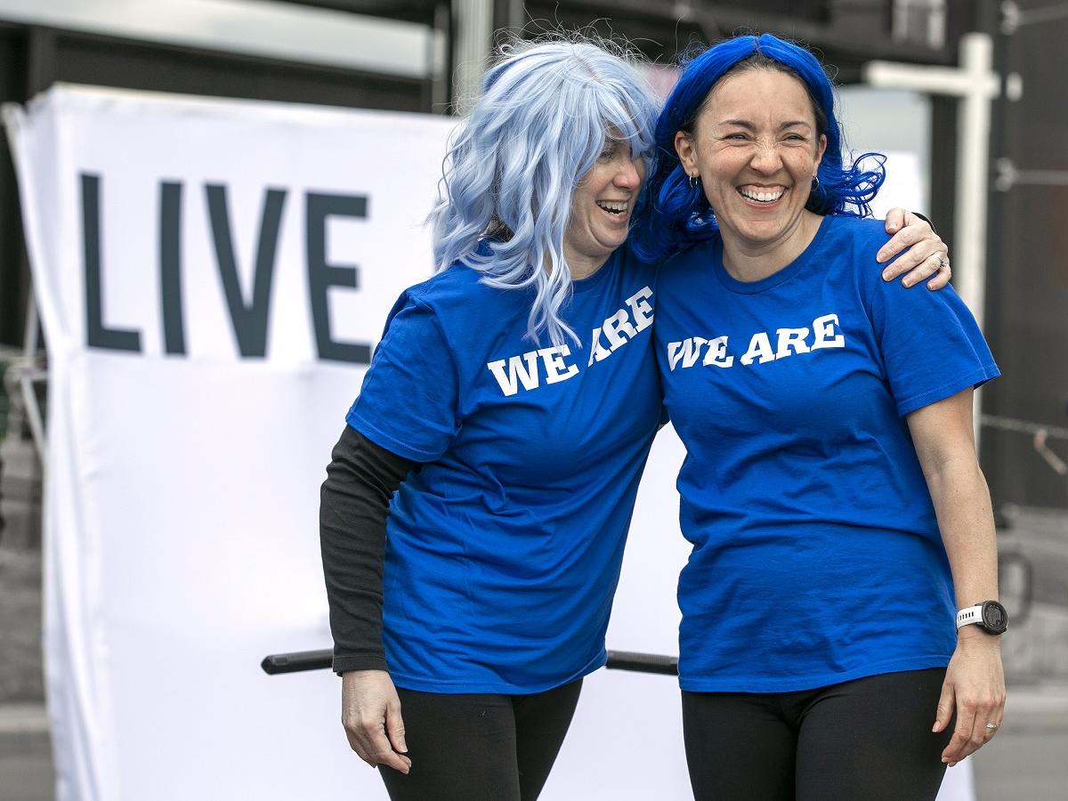 Two women stand arm-and-arm and laugh. They wear T-shirts that say "We Are."