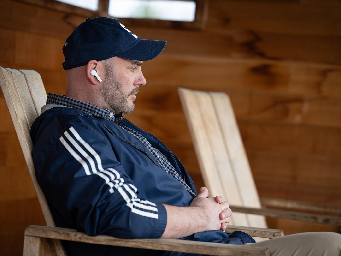 A man wearing a baseball hat gives a contemplative looks while he reclines in a wood chair.
