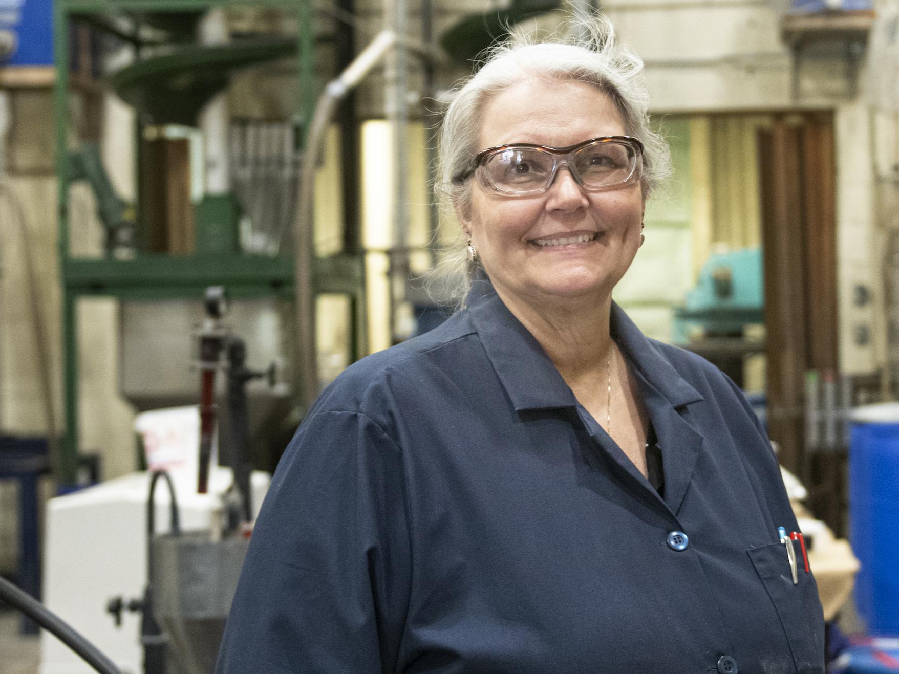 Woman in blue shirt and safety glasses in a lab looking at camera.
