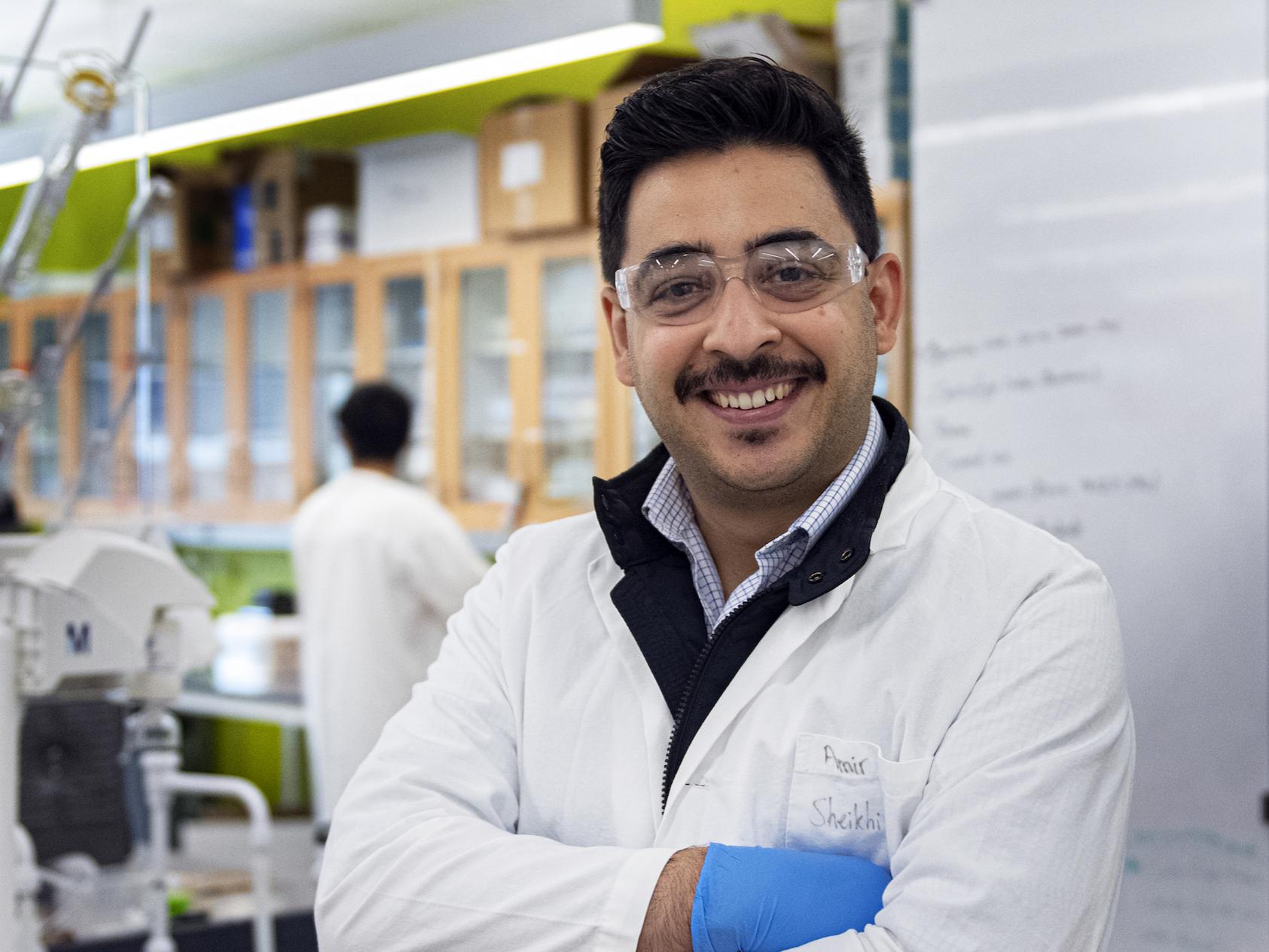 Smiling dark-haired man with mustache and white lab coat smiling with arms folded.