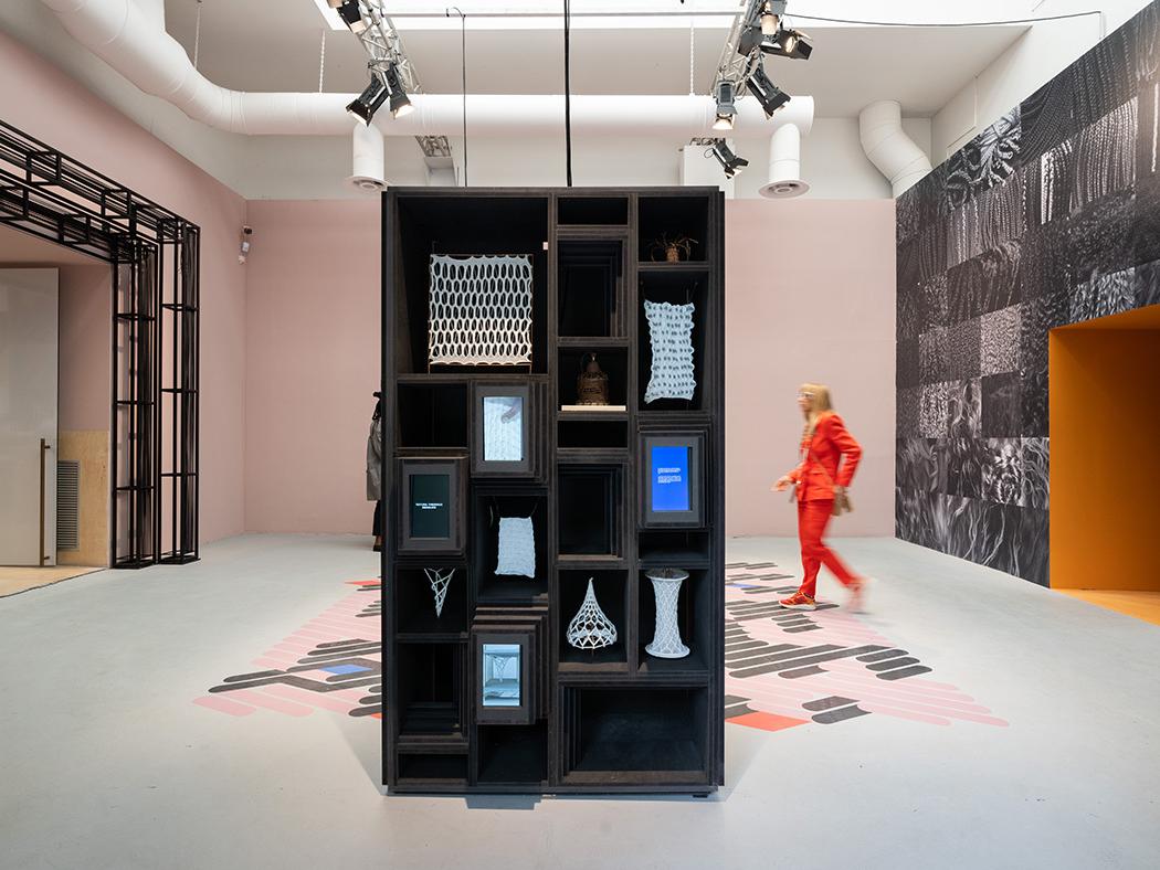 The Hair Salon exhibit picturing a work station featuring braided textiles and a person in an orange suite walking past.