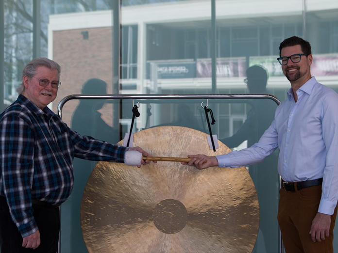 Two men in front of gong, one is handing the other a mallet