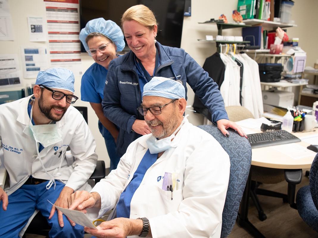 A man wearing glasses and a lab coat is seated in the foreground holding a photo. A man sits on the left, also is wearing glasses and a lab coat. Both men wear a mask around their neck and a surgical cap on their head. Two women stand behind them. The woman on the left is wearing a surgical bouffant. The woman on the right is wearing a sweat jacket and has a stethoscope around her neck. A keyboard sits on the table to the right. In the background are posters on the wall, a TV monitor and shelves.