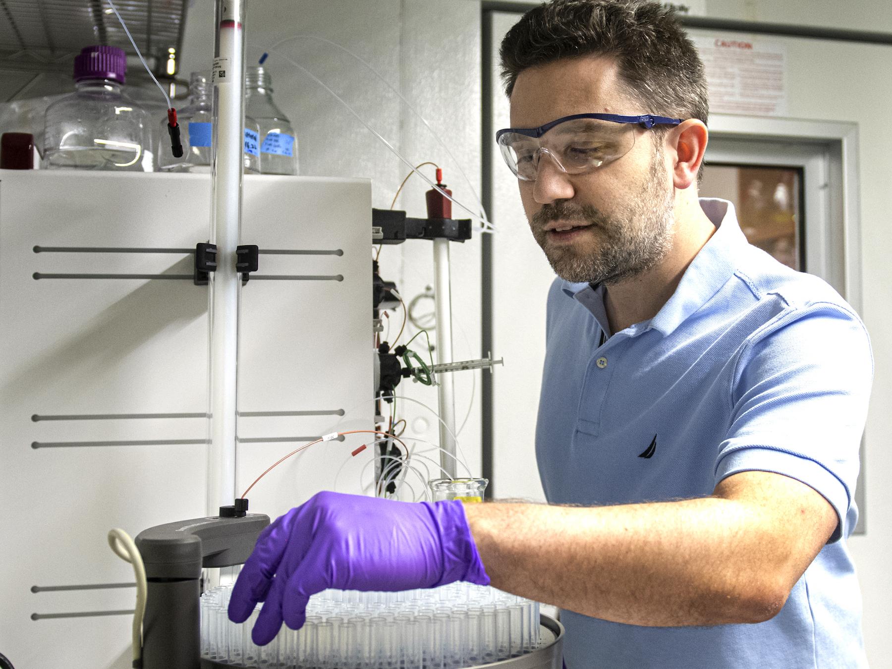 Man in blue polo shirt manipulates lab equipment with purple-gloved hand.