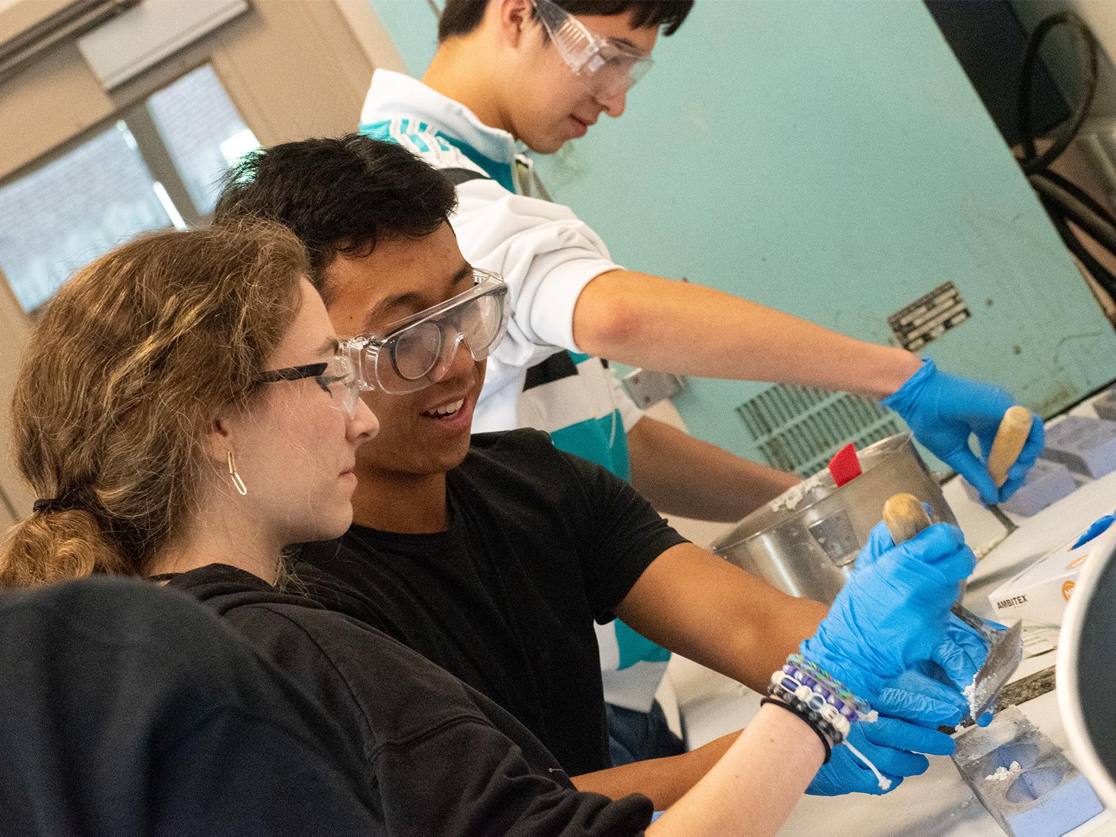 Students wearing safety goggles and blue gloves work with concrete during a civil engineering workshop