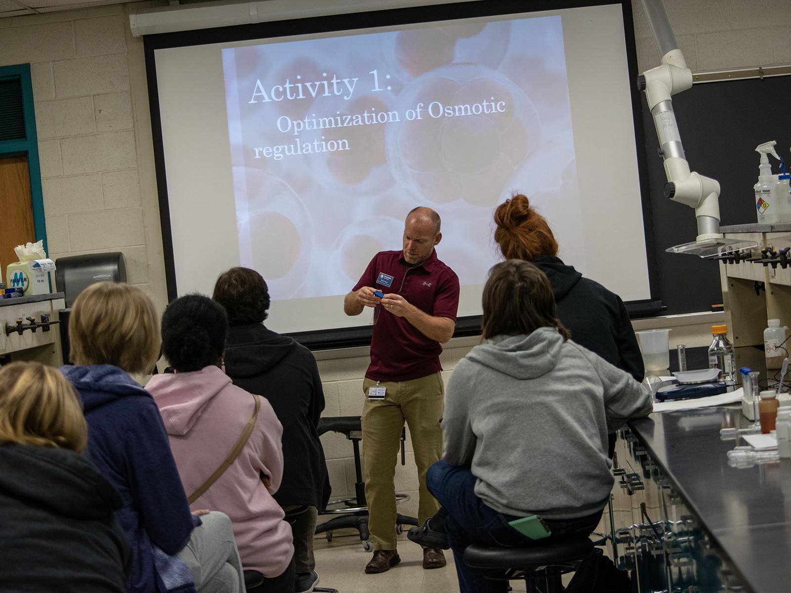 Students sit at lab tables while a man at the front of the room gives instructions during a biology workshop