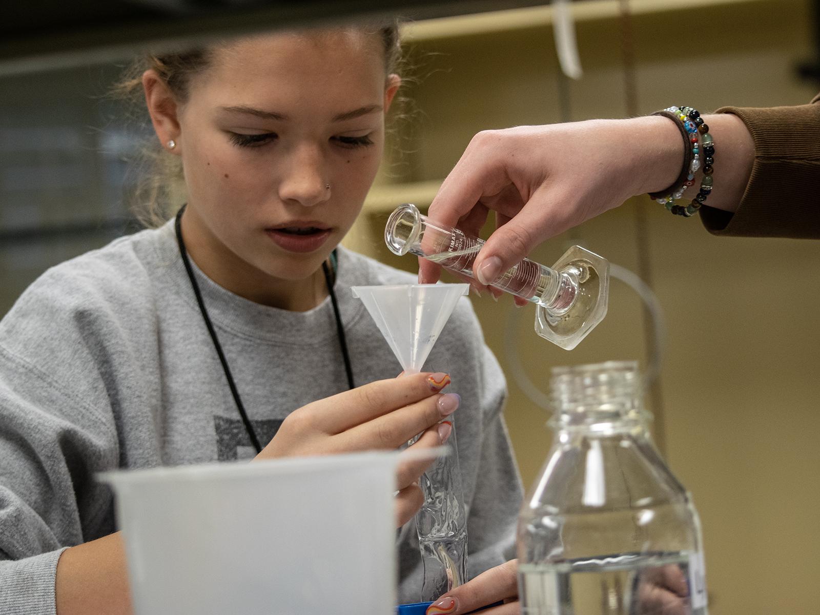 A high school student holds a funnel while another student pours liquid into a container.