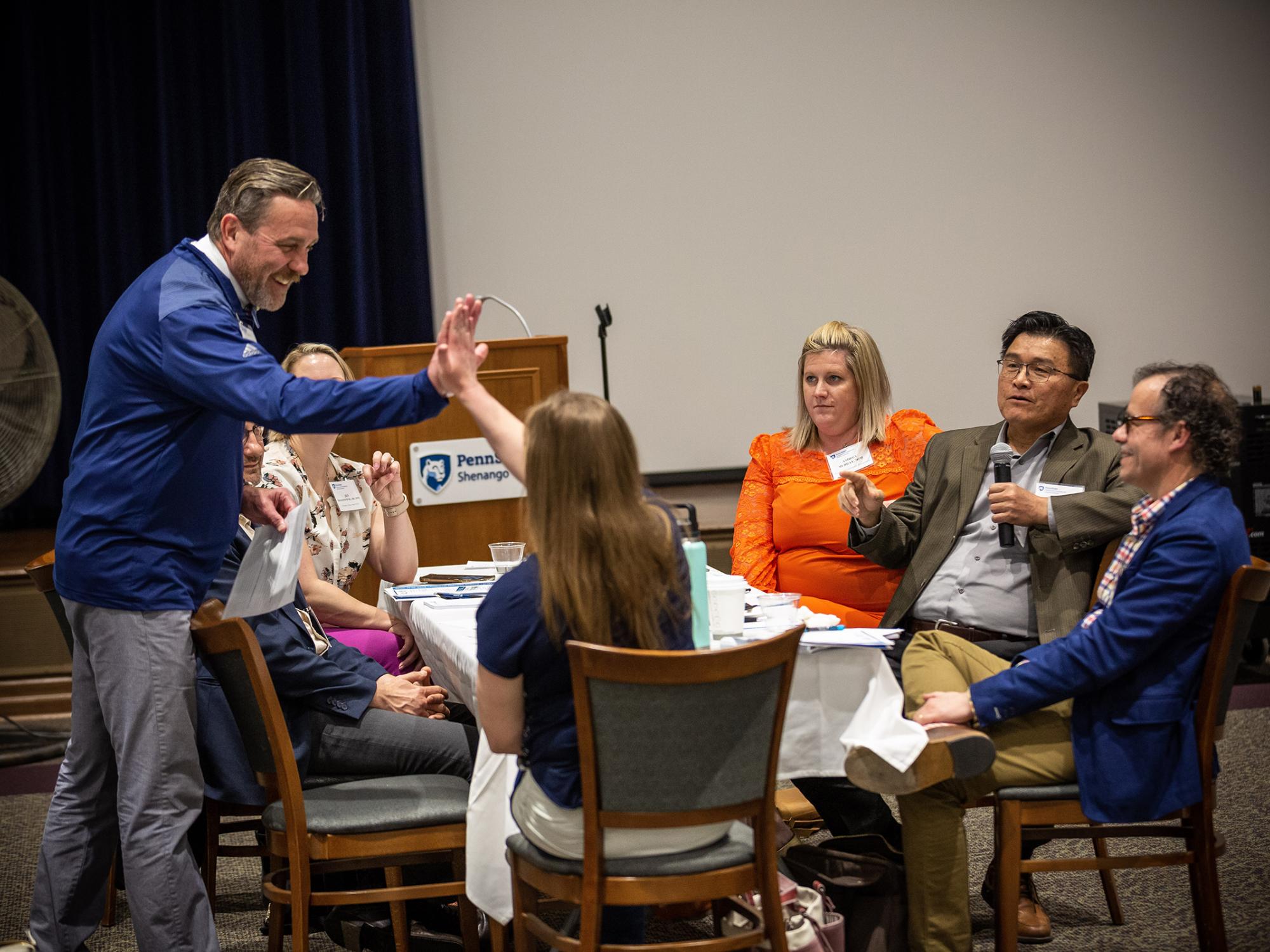 Two people hi-five each other at a table of people at a conference.