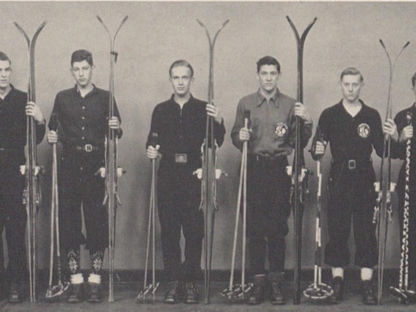 A black and white photo of male students holding skis and poles