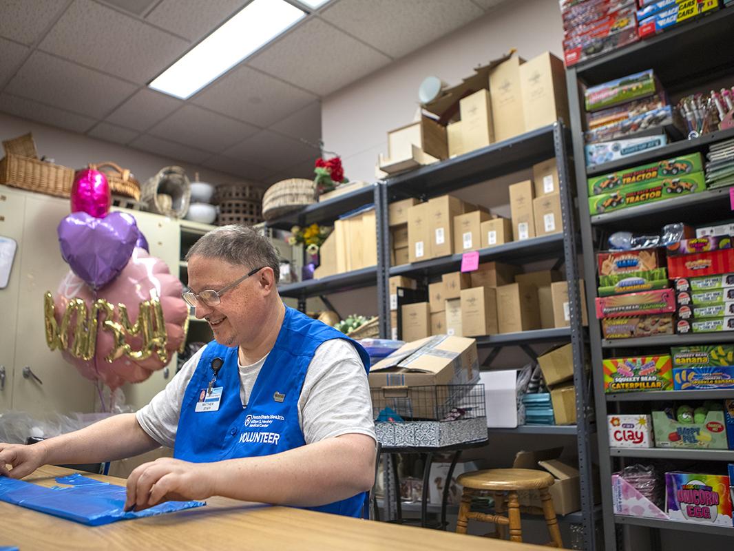 A man wearing a blue hospital volunteer vest is seated at a table organizing bags in a hospital gift shop stock room.