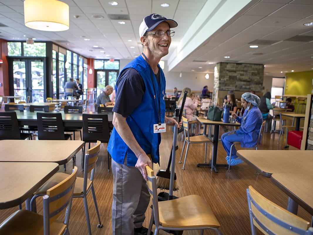 A man wearing a blue hospital volunteer vest sweeps a floor in hospital cafeteria.