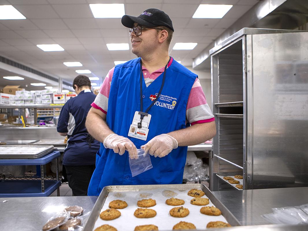 A man wearing a blue hospital volunteer vest sorts cookie in a hospital kitchen.