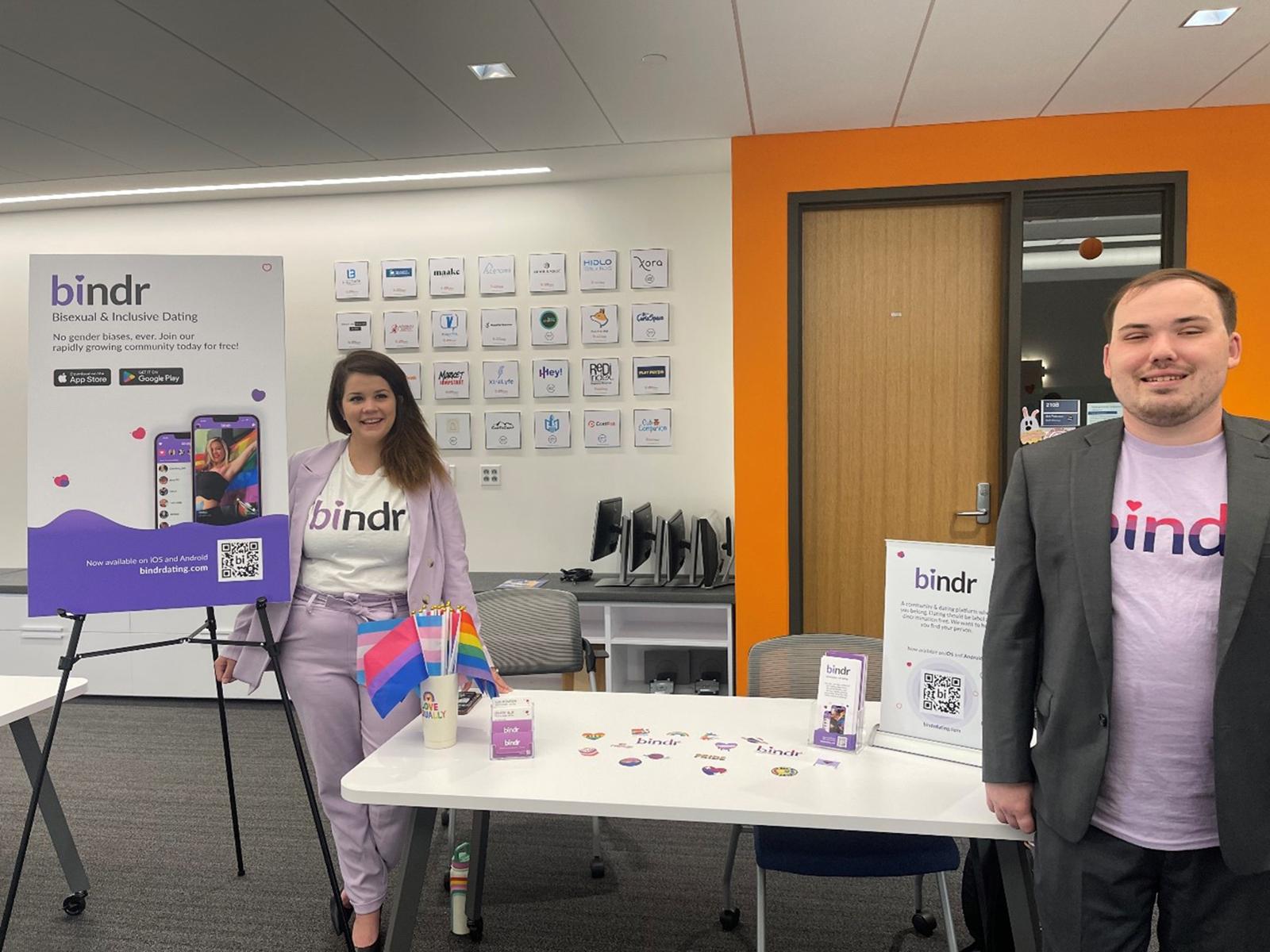 Two people stand in front of display boards at tables promoting an app called Bindr