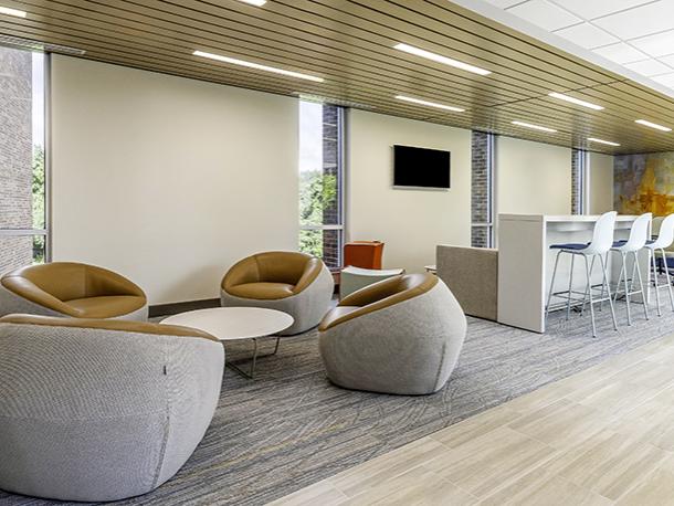 Comfortable chairs and a high table and chairs are part of a common area. There are large windows and at the far end of the area is an abstract painting in warm colors.