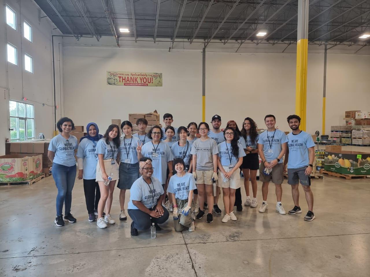 A large group of Penn Staters and international students in the Global Health Exchange Program stand in a large warehouse at the Central Pennsylvania Food Bank in Harrisburg, helping to pack food boxes.
