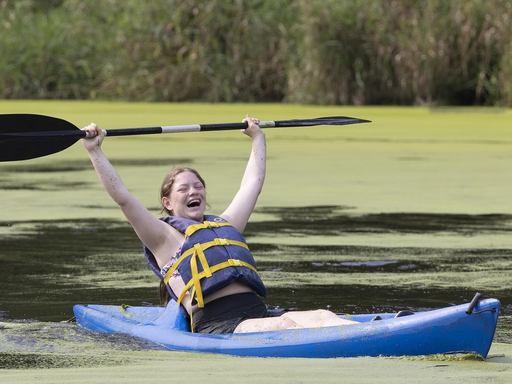 A girl in a kayak raises her paddle over her head and laughs.