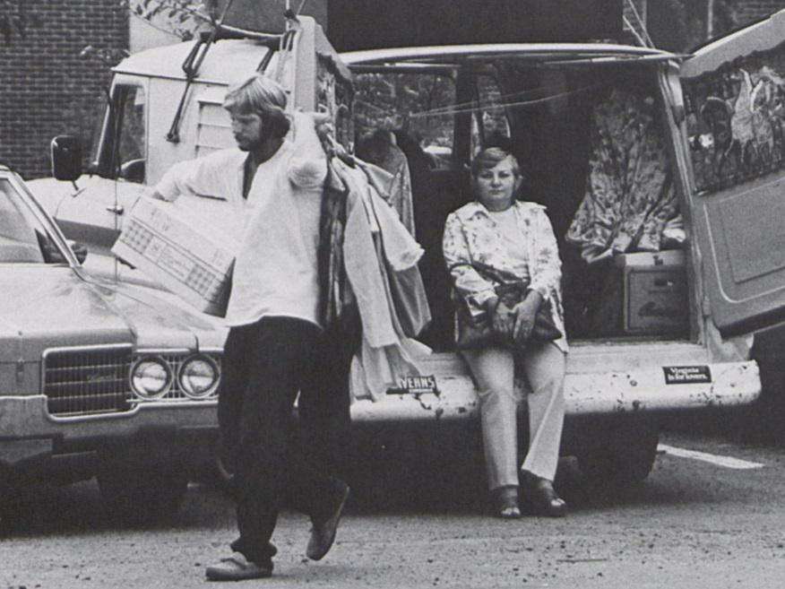 A male student carries a box and a handful of clothing while an older woman rests in the background