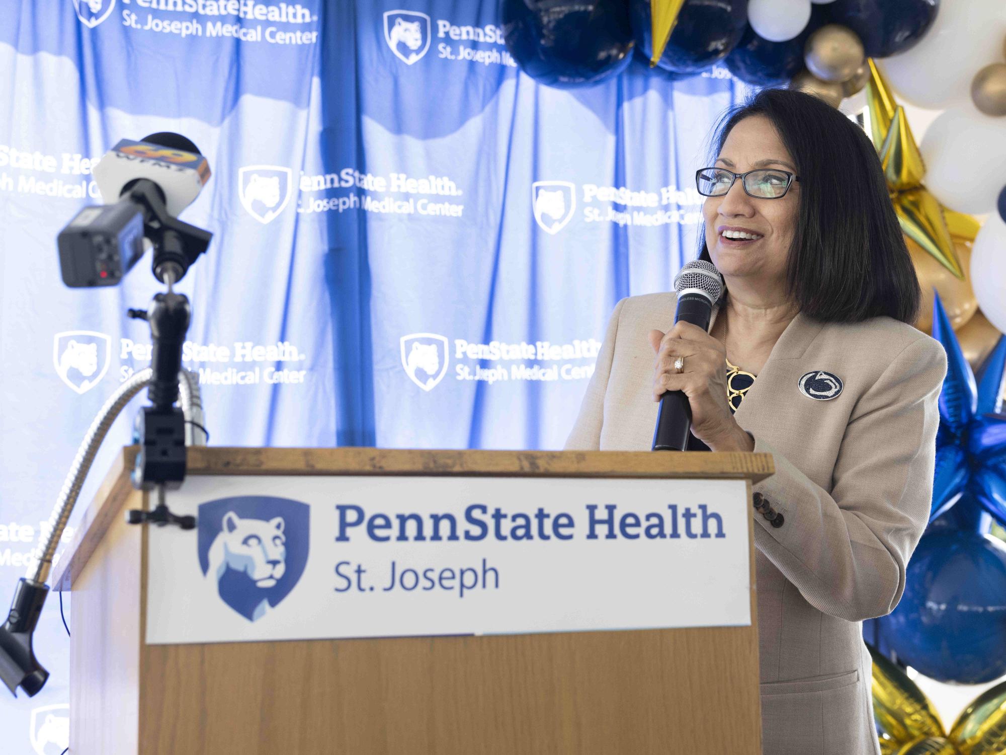 A woman in professional attire and a Penn State pin stands at a lectern with a microphone. The Penn State Health St. Joseph logo appears throughout the backdrop and on the lectern.