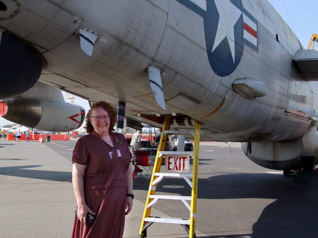 A woman stands beneath a World War II airplane. To her right, a ladder provides entrance to the plane. An EXIT sign hangs from the ladder. In the background between the woman and the ladder is a folding chair. People are visible walking on the airport tarmac behind her.