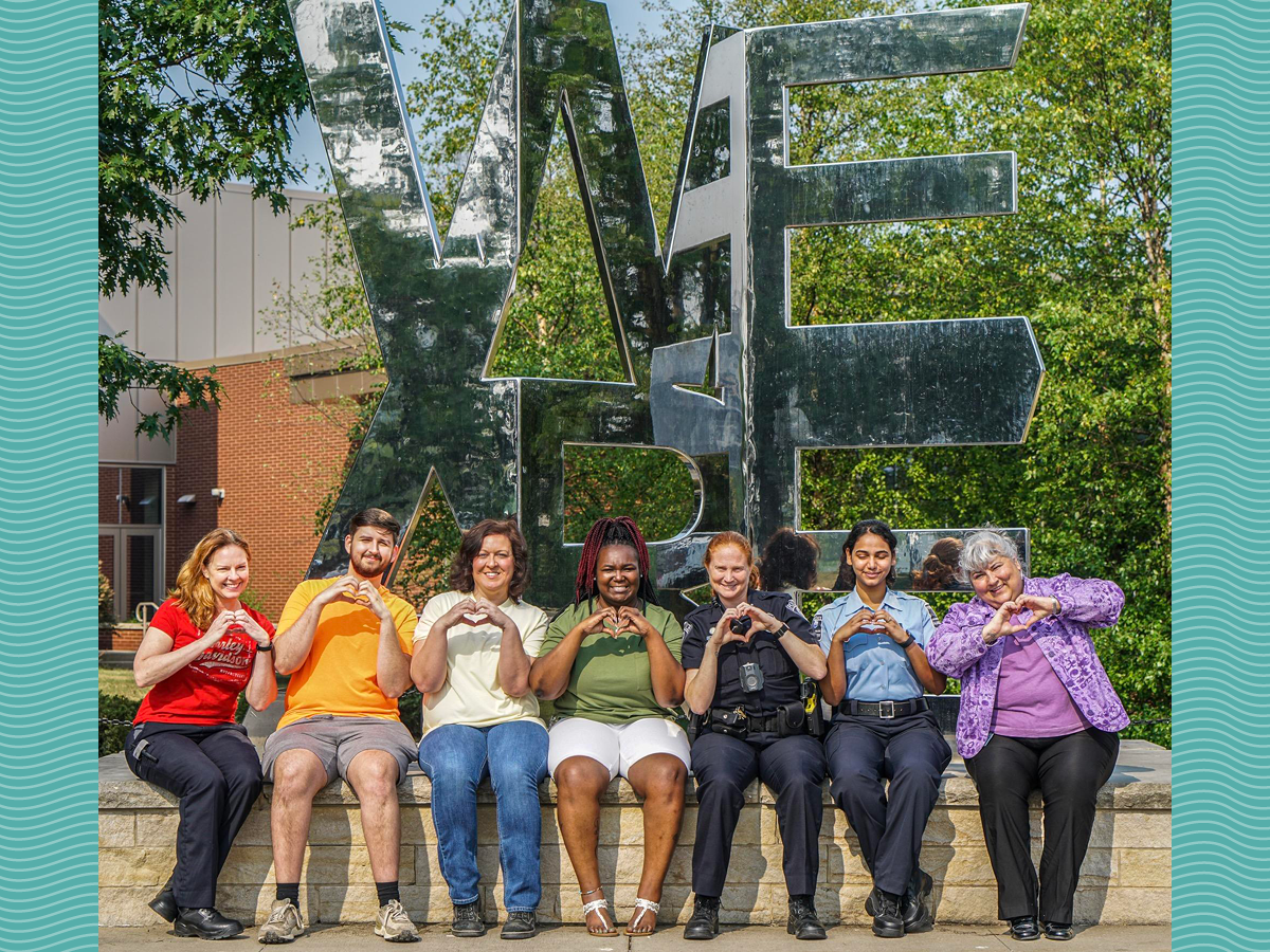 People in rainbow colored clothing sit in front of "We Are" statue with heart hands.