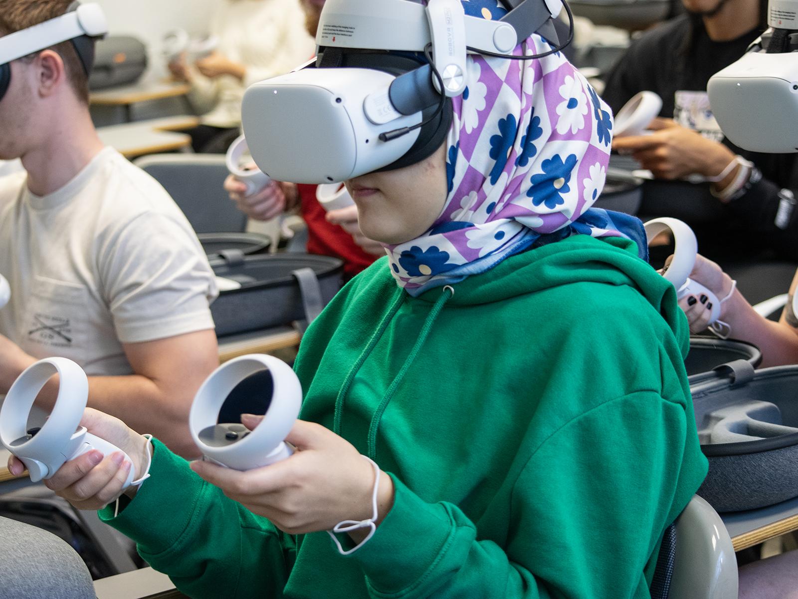 A student wears a virtual reality headset and holds a controller in each hand