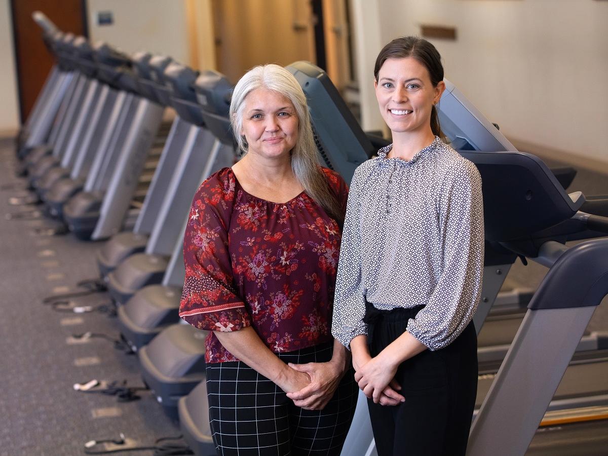 Two women stand in front of a row of treadmills. 