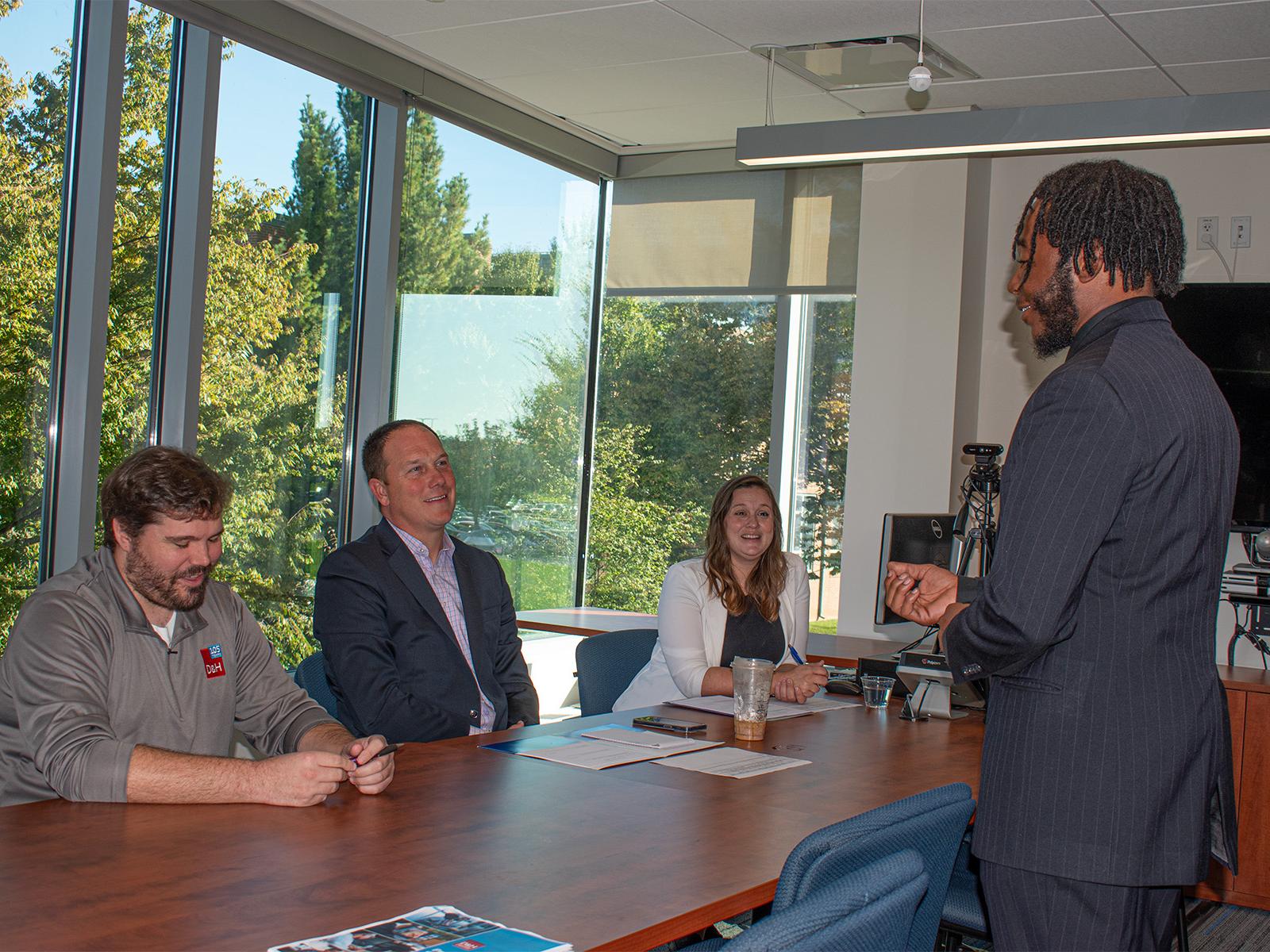 A student in a suit speaks before three company representatives sitting at a table