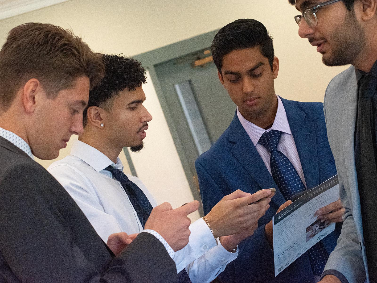 Four students stand together, looking at their phones and holding company brochures