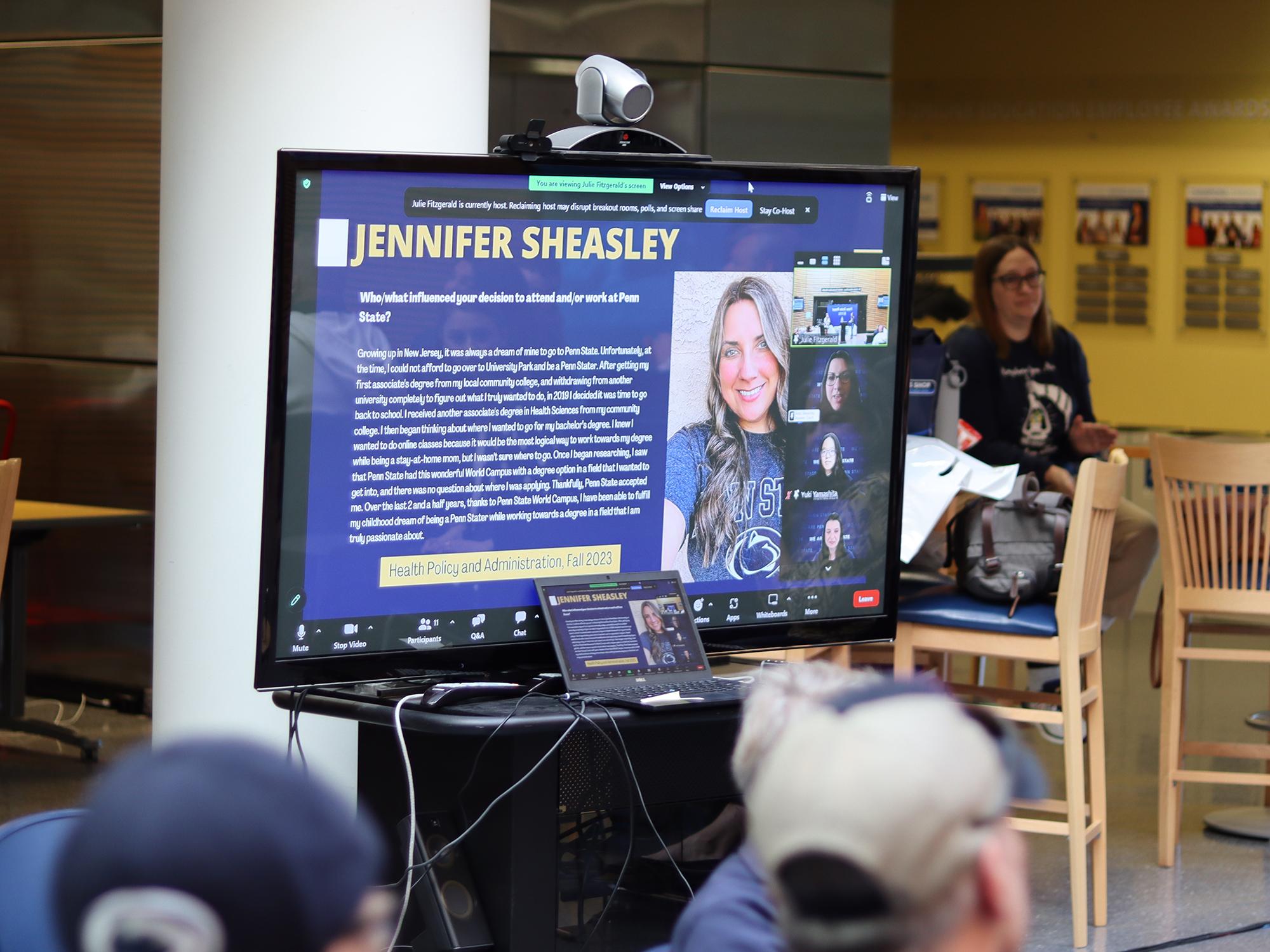A group of people watch a woman speak on a TV screen.