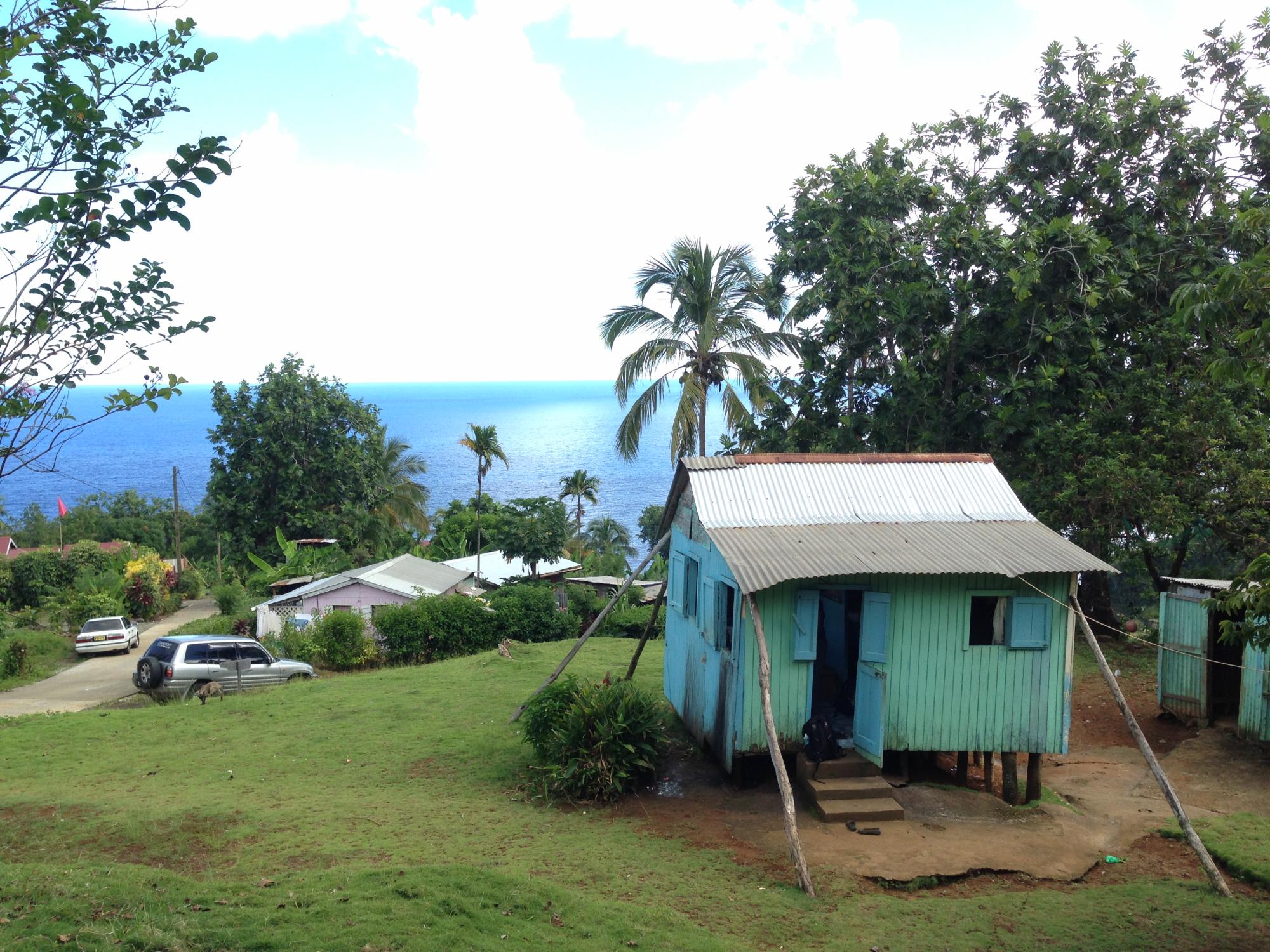 A blue house on a green hill. Other houses, trees and the ocean are visible in the background. 