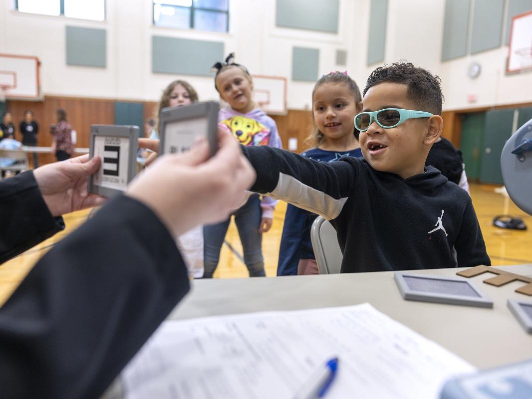 A little boy in glasses points at an image held by an adult. A line of children stand behind him.