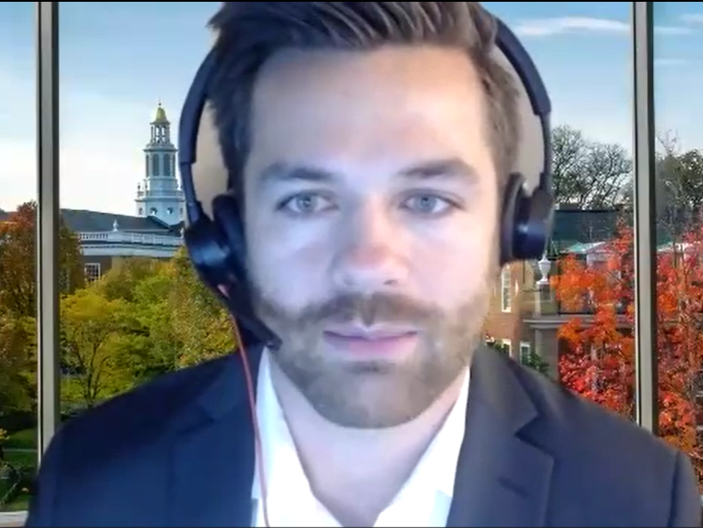 Tim Kluska, wearing headphones, gives a speech from his computer, wearing a suit and tie with the campus of Harvard in the background