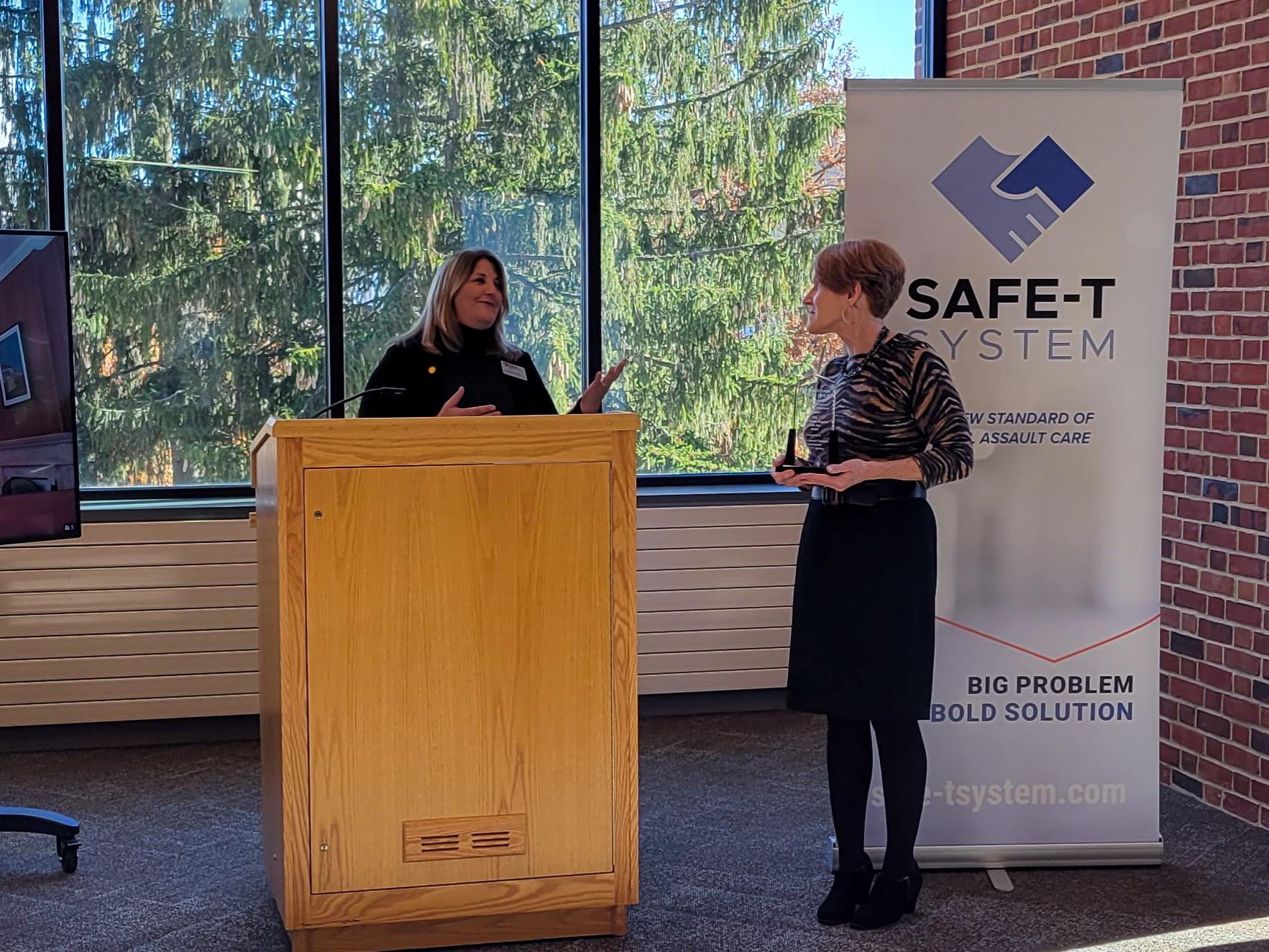 Cynthia Bittner, a woman in a black sweater behind a podium, accepts the award from Lisa Davis, a woman in a patterned shirt and black skirt.