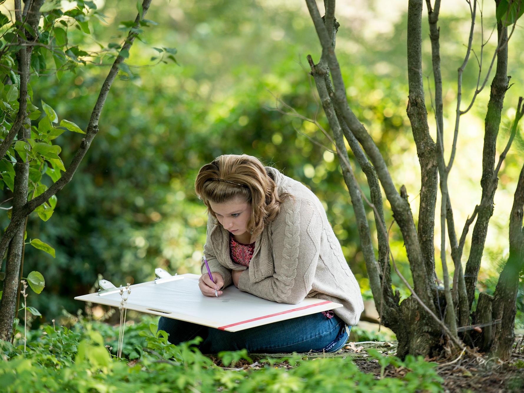 A student sits in front of a small plant and studies it with a large art book on her knees.
