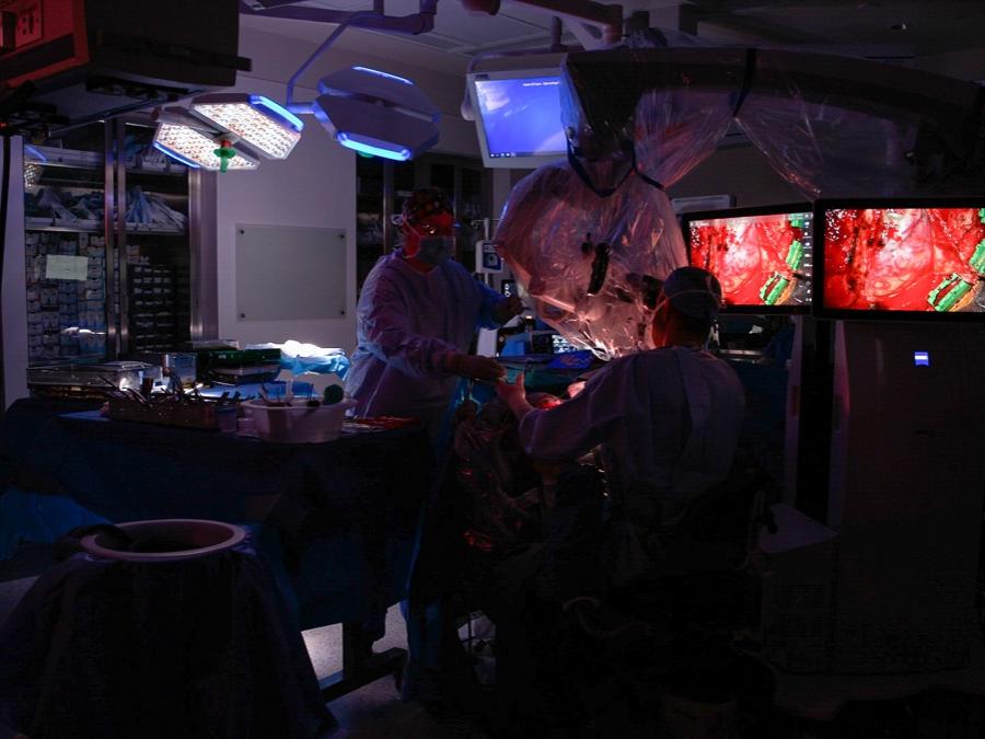 Interior of a darkened operating theater. Two people in scrubs stand before close-up video images of a brain.