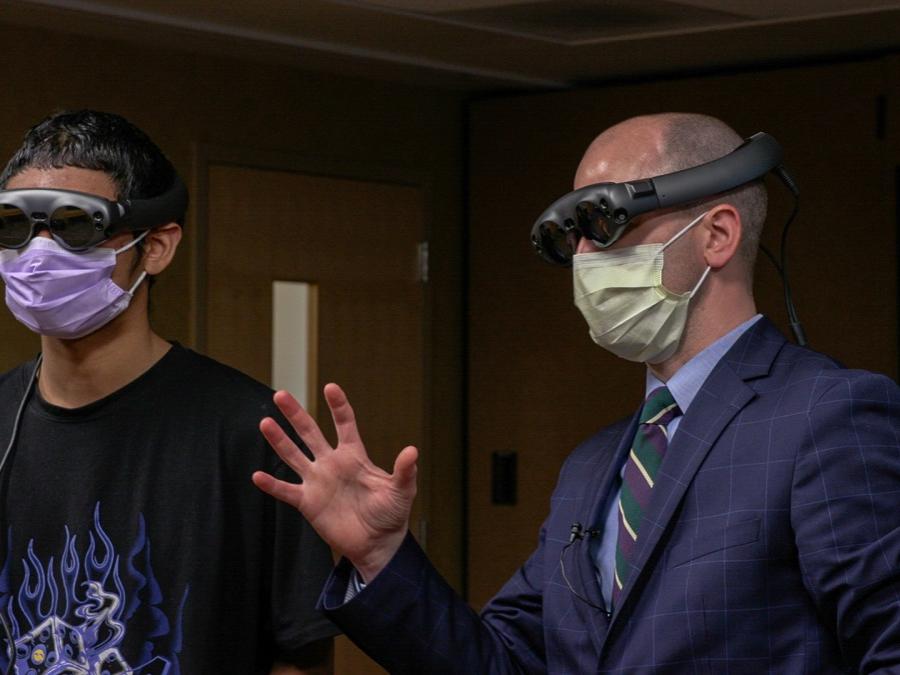 A boy in a T-shirt stands next to a man in a suit. Both wear goggles. The man is gesturing.