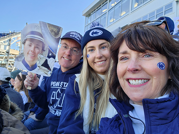 Three people sit in the stands at Beaver Stadium wearing Penn State attire - One is holding a photo of their son.