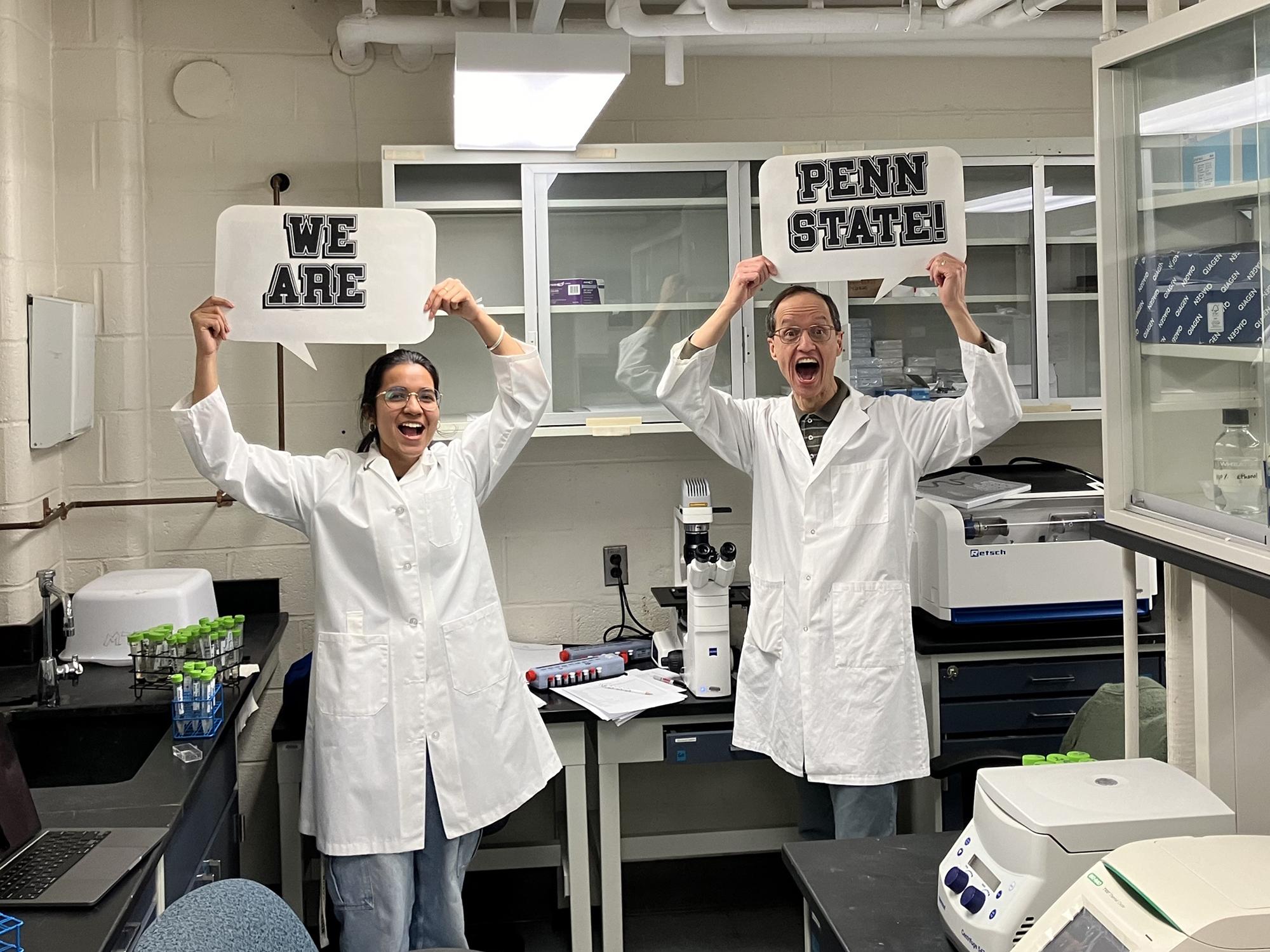 Two researchers wearing lab coats hold up signs saying "We Are" and "Penn State"