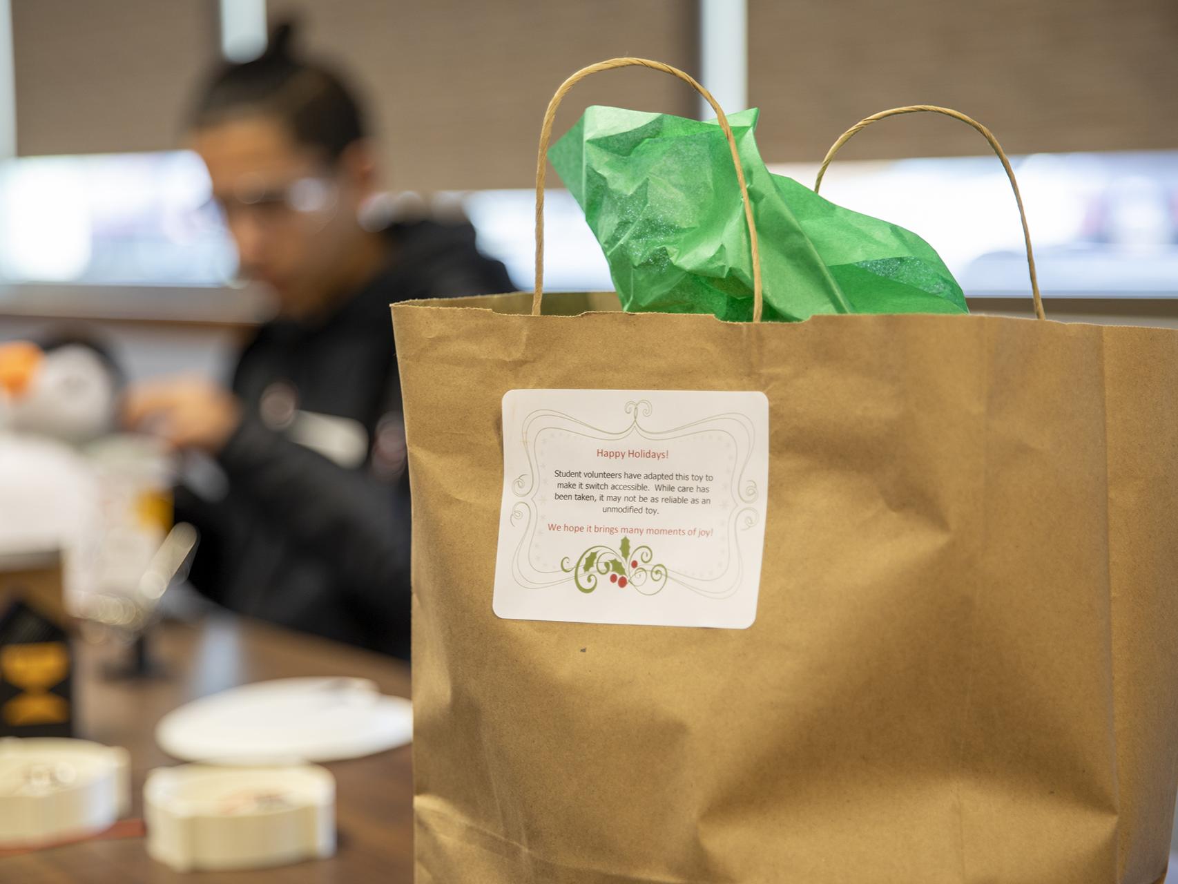 A gift bag with green tissue paper sits on a table in the foreground with a high school student assembling a button in the background.