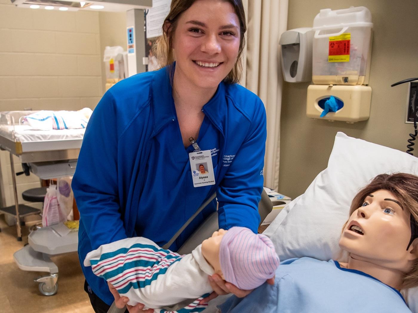 A nursing student wearing blue holds a baby doll while standing next to a bed containing a manikin.