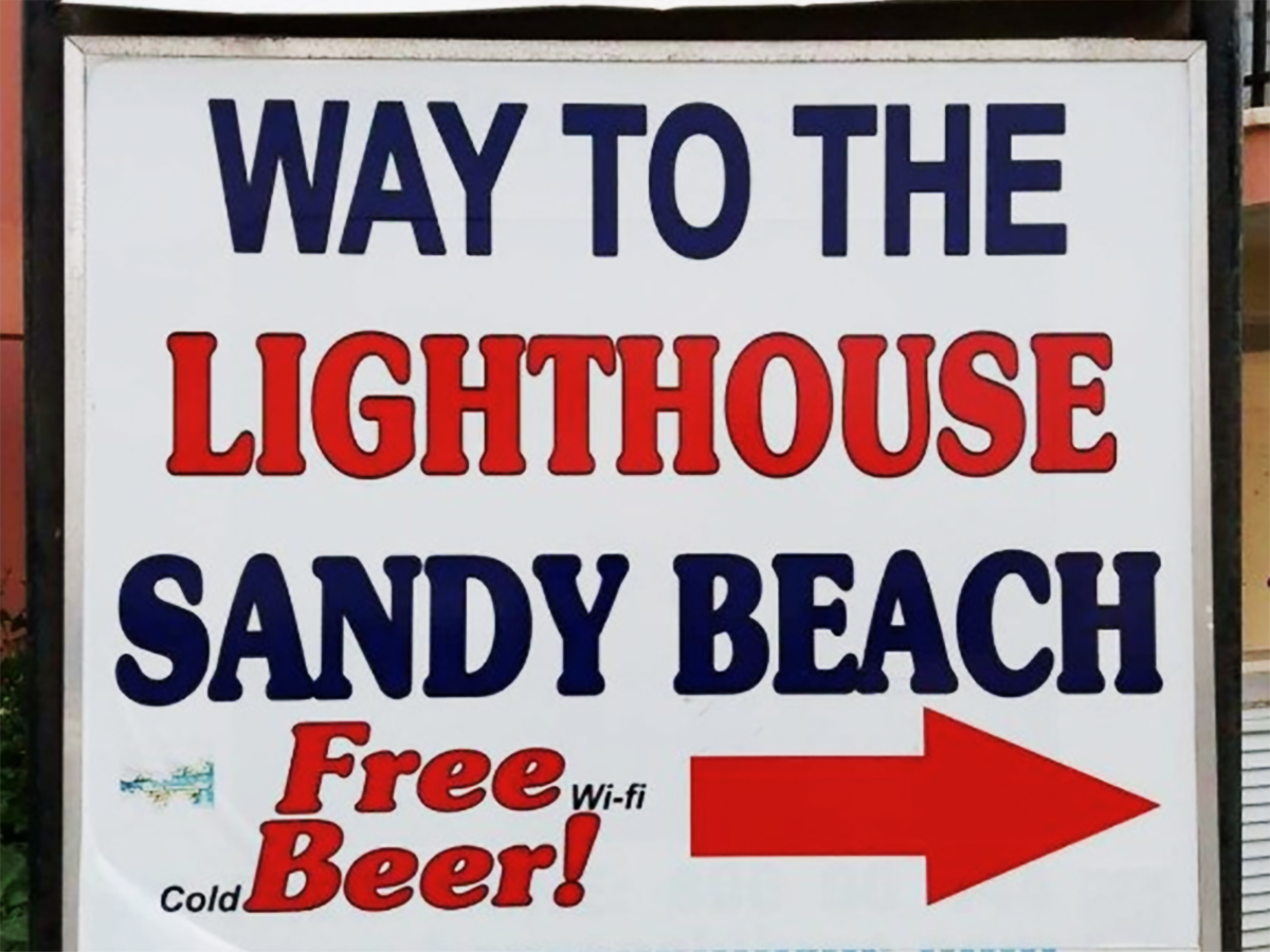 A sign reading "Way to the Lighthouse Sandy Beach, Free Wi-fi Cold Beer!" and an arrow pointing right
