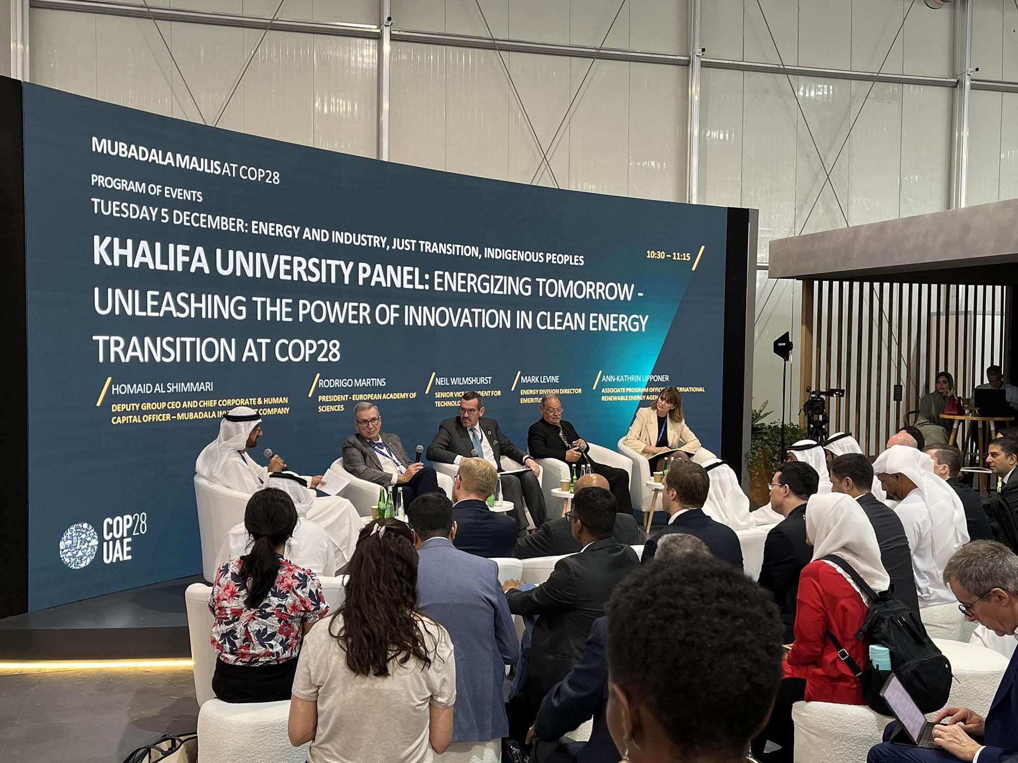 Five people on a stage discuss a topic while an audience looks on