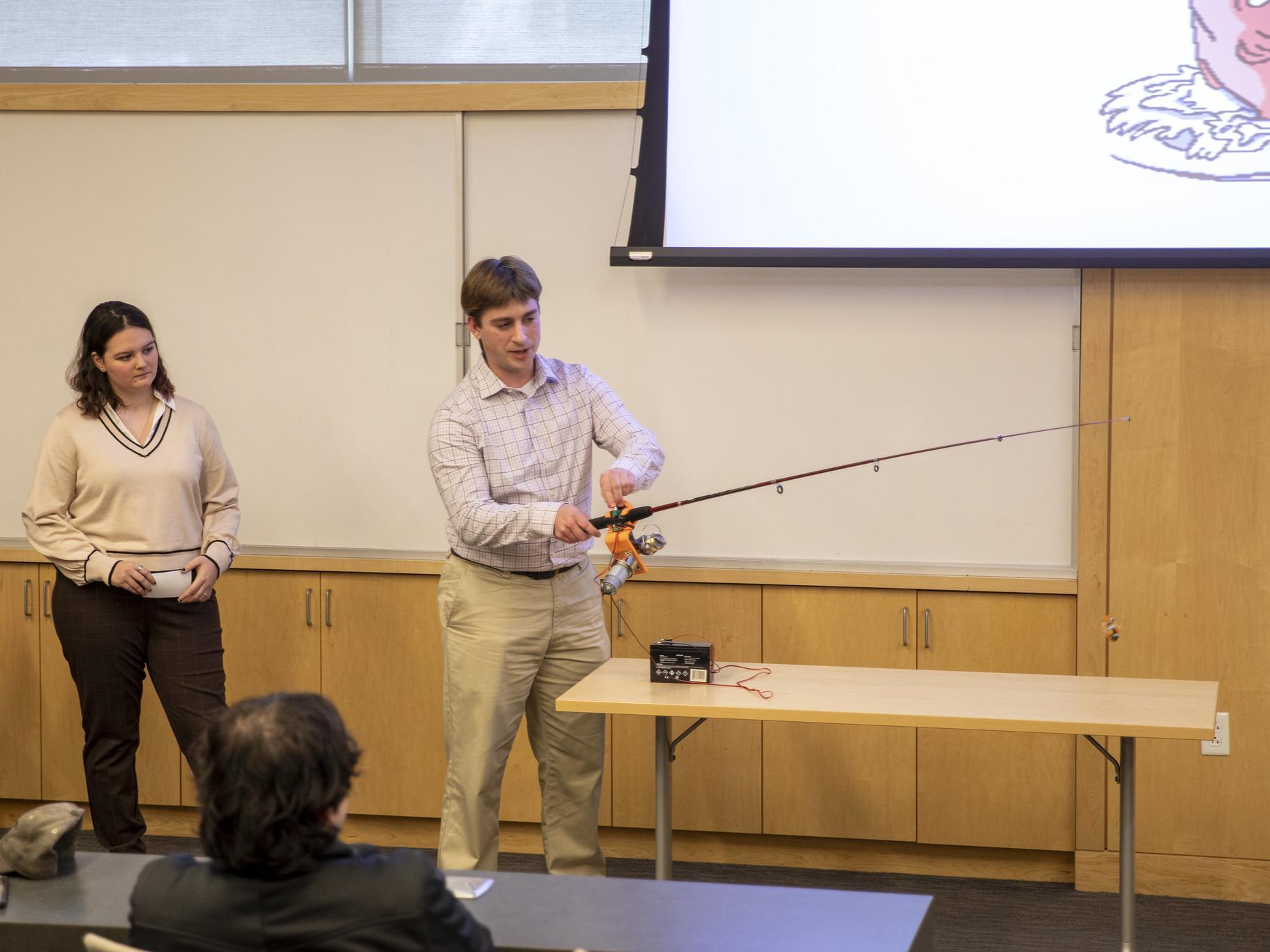 Male student holds adaptive fishing rod to demonstrate to group of attendees