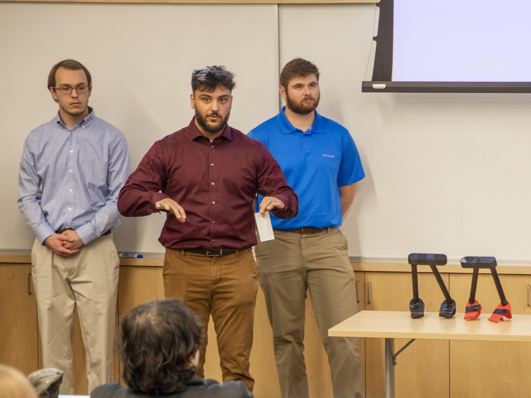 Three male students present portable assistive toilet handles