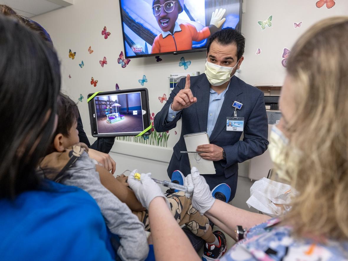 A man in a surgical mask and a sport jacket holds up an index finger while talking to three people in a hospital room.