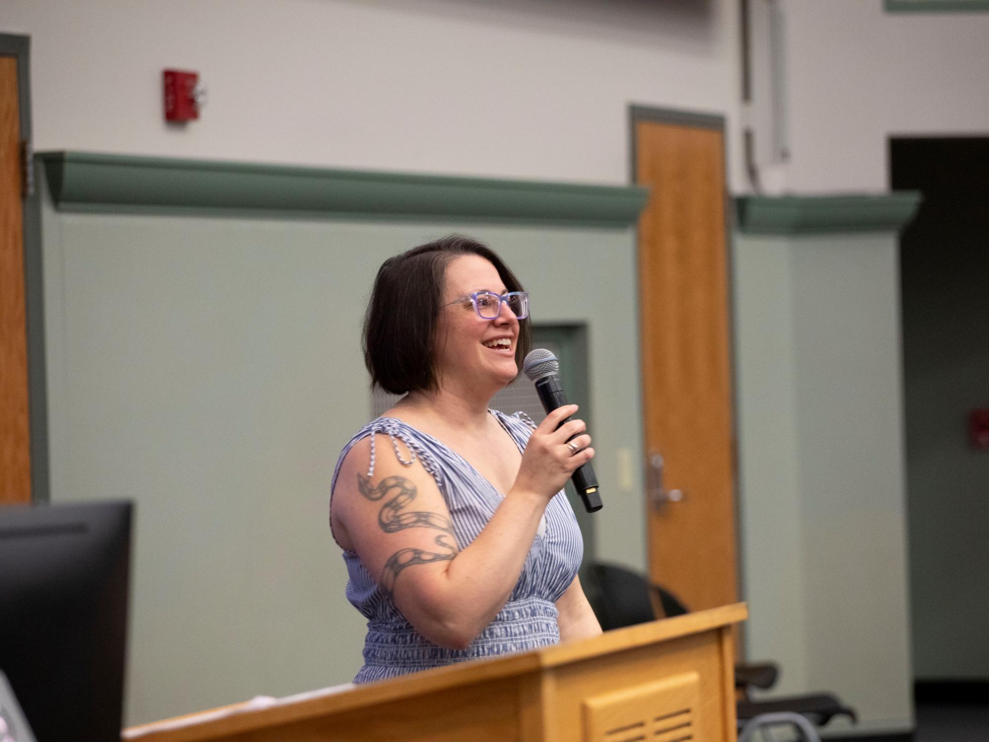 teacher with dark hair and glasses speaks into microphone at podium